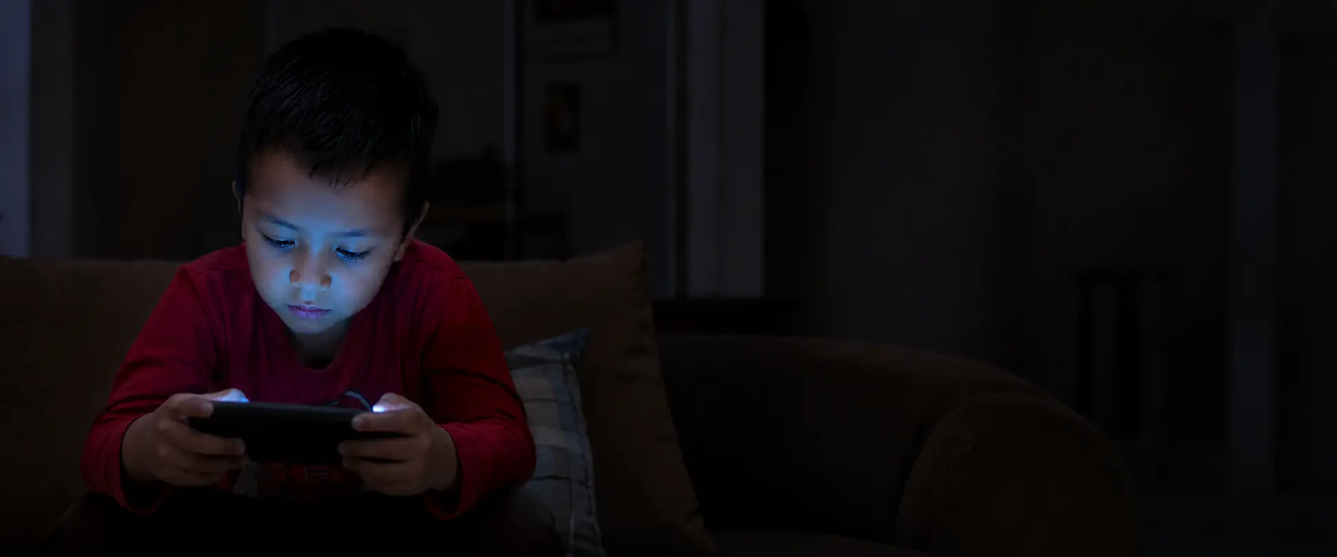 Niño absorto en su celular, iluminado únicamente por la pantalla en una habitación oscura